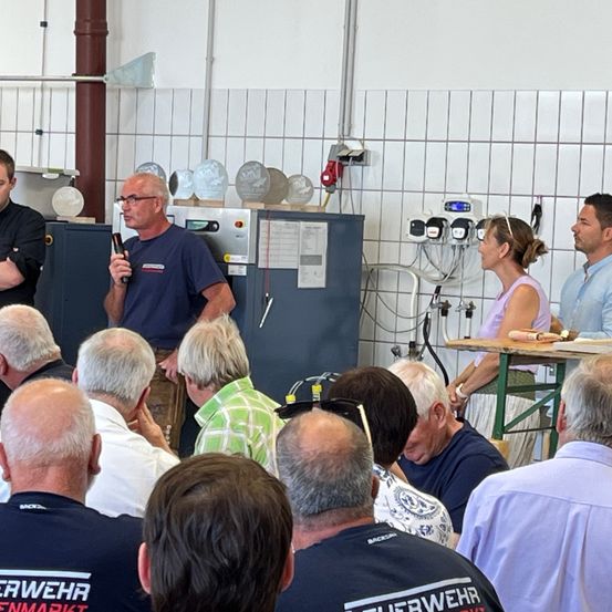 Eine Gruppe von Menschen ist in einem Raum versammelt, möglicherweise für eine Besprechung. Ein erwachsener Mann mit Brille steht vor einer Gruppe und hält ein Mikrofon. Hinter ihm steht ein großer grauer Schrank mit angehefteten Papieren. Eine Frau in einem lila Shirt schaut ihn an.