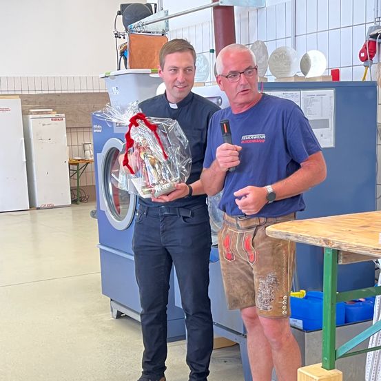 Zwei Männer stehen in einem Raum, einer hält ein in Plastik eingewickeltes Geschenk, der andere ein Werkzeug. Sie sind in der Nähe einer Waschmaschine und eines Holztisches.