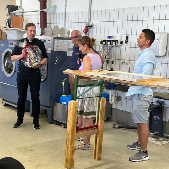 Vier Personen stehen um einen Holztisch in einem Hauswirtschaftsraum, ein Mann hält ein Geschenk. Eine Waschmaschine steht hinter ihnen.