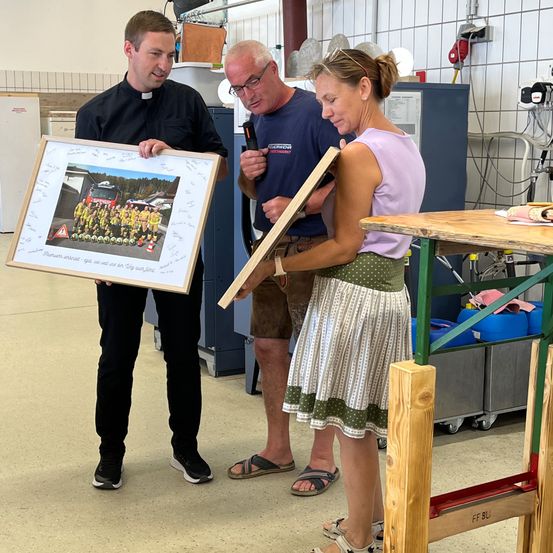 Drei Menschen stehen in einem Raum und halten einen Fotorahmen und eine Holzplatte. Ein Mann in einem schwarzen Hemd hält einen Fotorahmen, während eine Frau in einem Rock eine Holzplatte hält. Der andere Mann hält eine Kamera. Dahinter steht ein Holztisch mit Gegenständen darauf.