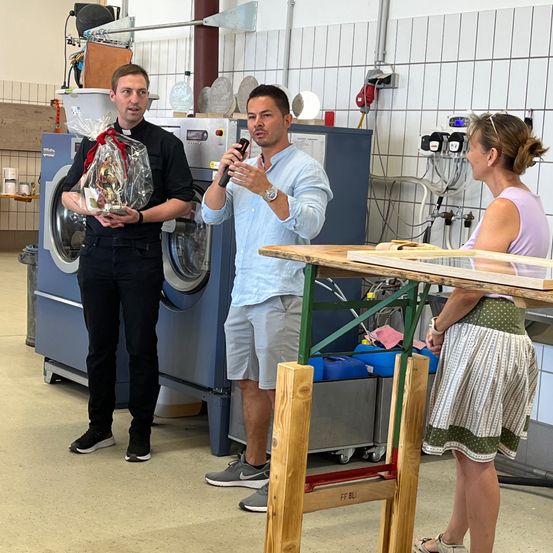 Drei Personen stehen in einem Raum mit einem Holztisch und Waschmaschinen. Ein Mann hält einen Geschenkkorb und spricht in ein Mikrofon.