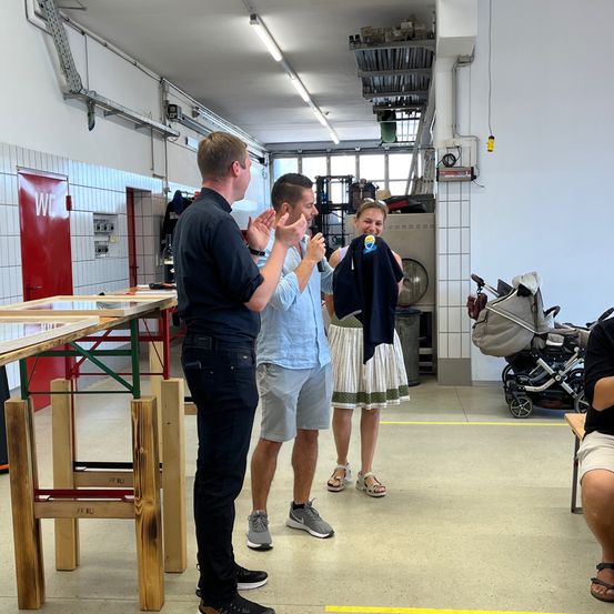 Drei Personen befinden sich in einer Werkstatt. Eine Frau hält einen blauen Mantel. Ein Mann klatscht. Ein anderer Mann lächelt. Ein Kinderwagen und ein Holztisch sind im Hintergrund.
