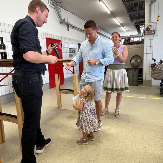 Eine Familie ist in einer Werkstatt. Ein Mann hält ein Werkzeug, ein Kind schaut auf, eine Frau klatscht und ein Kinderwagen steht in der Nähe.