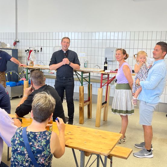 Eine Gruppe von Menschen versammelt sich in einem Raum, ein Mann mit einem Mikrofon spricht, während andere zuhören. Eine Frau mit einem Kind steht in der Nähe. Tische und Bänke sind vorhanden, und es gibt Küchengeräte an der Wand.