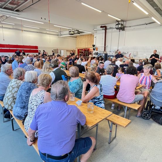 Eine Gruppe von Menschen sitzt auf Bänken in einem Raum, wahrscheinlich in einer Feuerwache. Einige tragen Hüte und Turnschuhe. Einige von ihnen schauen den Mann an, der vor den Tischen steht. Einige von ihnen halten Gläser. Auf den Tischen stehen Flaschen, Tassen und Teller. Die Decke hat ein paar Lichter.