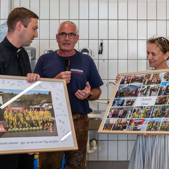 Ein Mann in einem blauen T-Shirt mit der Aufschrift Feuerwehr Eurochmarkt spricht in ein Mikrofon. Neben ihm hält ein Mann in einem schwarzen T-Shirt ein gerahmtes Foto. Eine Frau hält eine Collage von Feuerwehrfotos.