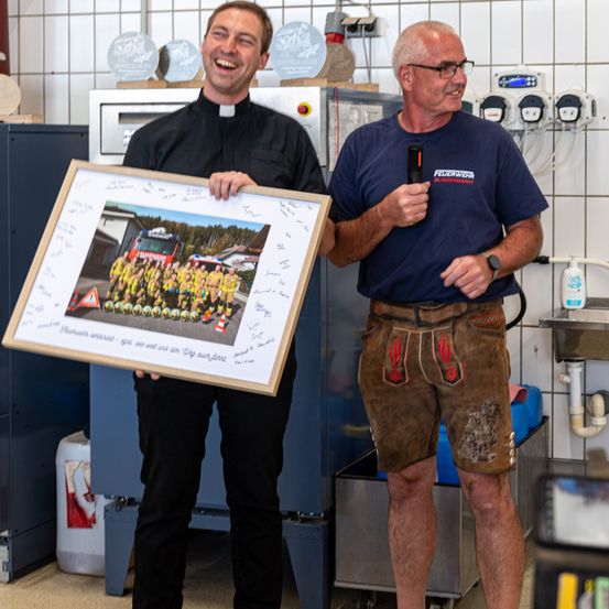 Ein Mann in einem schwarzen Klerikerkragen, der ein gerahmtes Bild einer Feuerwehr hält, steht neben einem anderen Mann in traditioneller bayerischer Kleidung, der ein Mikrofon hält.