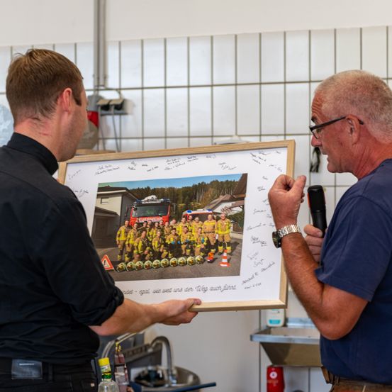 Zwei Männer betrachten ein gerahmtes Foto von Feuerwehrleuten, wobei einer ein Mikrofon hält und der andere auf das Bild zeigt. Sie stehen in einem Raum mit gefliesten Wänden und einem Waschbecken.