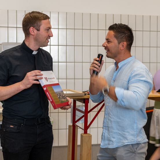 Zwei Männer stehen in einem Raum, einer hält ein Buch und der andere ein Mikrofon. Der Mann mit dem Buch scheint zu präsentieren oder zu sprechen, während der andere Mann aufmerksam zuhört. Hinter ihnen ist eine geflieste Wand.