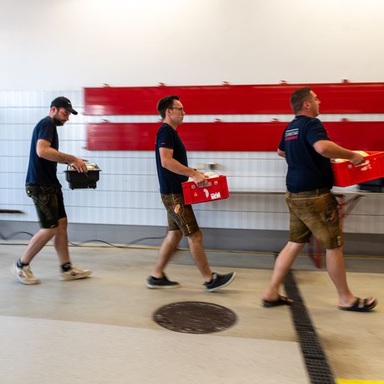 Drei Männer gehen mit roten Kisten in einem Raum. Sie tragen blaue Hemden und Shorts. Der Raum hat eine weiße und eine rote Wand.