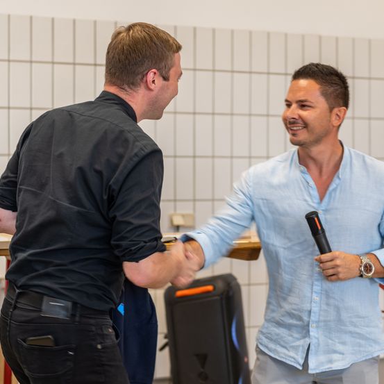Zwei Männer schütteln sich die Hände in einem Raum. Der Mann auf der rechten Seite hält ein Mikrofon. Beide lächeln. Ein Lautsprecher steht hinter ihnen. Die Wand ist gefliest.