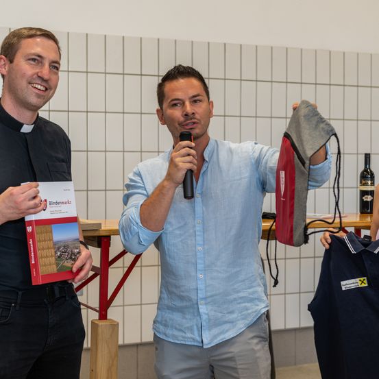 Zwei Männer stehen nebeneinander. Einer hält ein Buch, der andere ein Mikrofon. Sie lächeln beide. Dahinter ist eine weiß geflieste Wand.