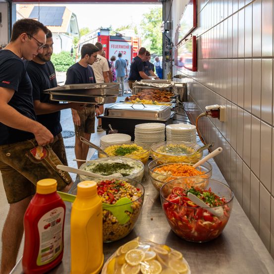 Freiwillige servieren Essen in einer Feuerwache. Mehrere Salate und Saucen stehen auf dem Tisch, mit einem Feuerwehrwagen im Hintergrund.