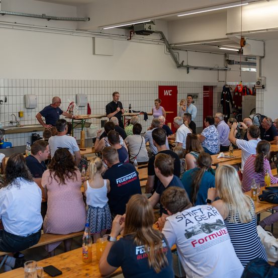 Eine Gruppe von Menschen sitzt an Tischen in einem Saal. Einige Leute stehen und sprechen davor. Ein Mann steht mit einem Mikrofon. Verschiedene Gegenstände sind auf den Tischen. Ein Waschbecken mit einem Wasserhahn ist auf der linken Seite.