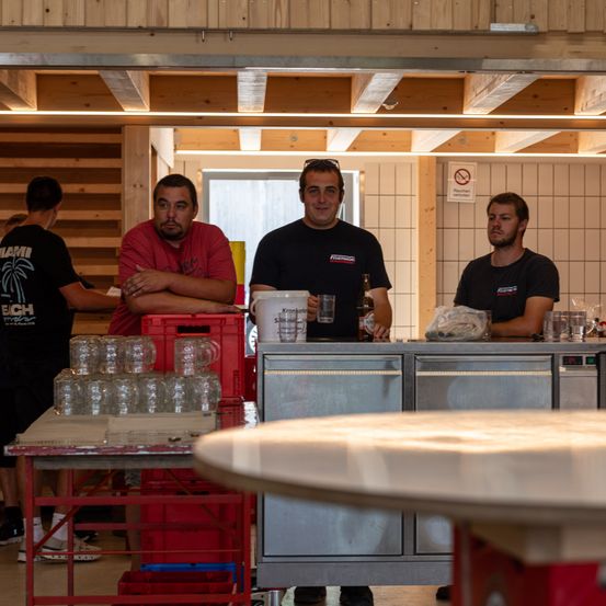 Vier Männer stehen hinter einer Bartheke in einem Raum mit Holzwänden. Zwei von ihnen tragen schwarze T-Shirts, eines mit einem Logo, und die anderen beiden sind in roten und schwarzen T-Shirts. Auf der Theke und vor ihnen stehen leere Gläser.
