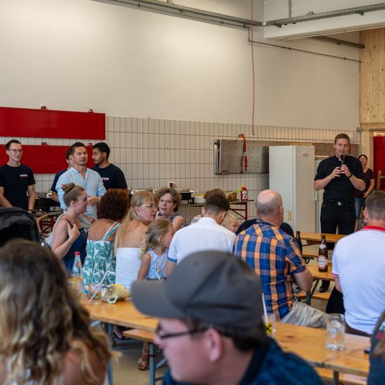 Eine Gruppe von Menschen in einem Raum mit einem Mann, der in ein Mikrofon spricht. Mehrere Tische mit Flaschen und Essen sind vorhanden. Eine Frau in einem schwarzen Shirt steht in der Nähe eines Kühlschranks.