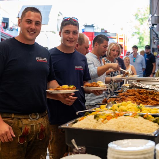 Zwei Männer in Uniformen servieren Essen bei einer Veranstaltung. Ein Buffet mit verschiedenen Gerichten steht vor ihnen. Menschen bedienen sich selbst. Ein Feuerwehrauto ist im Hintergrund zu sehen.