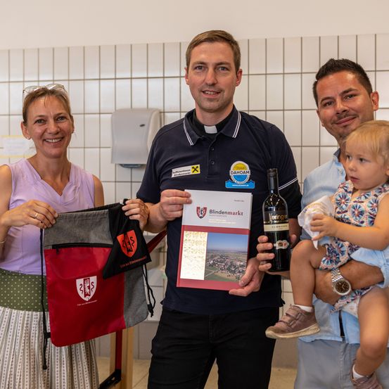 Vier Personen stehen lächelnd zusammen. Ein Mann hält eine Weinflasche und ein Buch mit dem Titel 'Blindenmarkt'. Eine Frau hält eine rote Tasche mit einem Logo. Ein anderer Mann hält ein Baby. Sie sind drinnen mit weißen gefliesten Wänden.