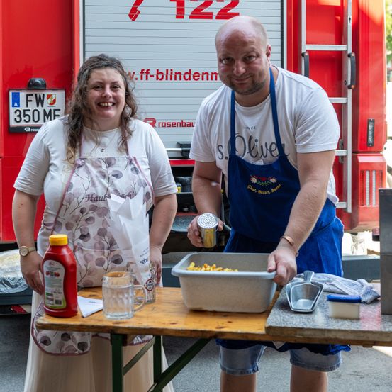 Ein Mann und eine Frau stehen lächelnd vor einem Feuerwehrwagen. Der Mann hält einen Messbecher und einen Behälter mit Essen, während die Frau eine Flasche Ketchup in der Hand hat. Sie tragen beide Schürzen.