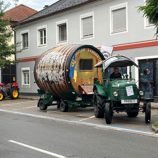 Ein grüner Traktor mit einem großen Fass-Anhänger vor einem Gebäude. Das Fass hat eine Flagge und Text darauf.