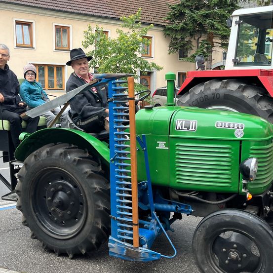 Drei Personen fahren auf einem grünen Traktor. Eine Person fährt, während zwei andere hinten sitzen. Der Traktor ist mit einem blauen und braunen Werkzeug ausgestattet. Gebäude und Autos sind im Hintergrund zu sehen.