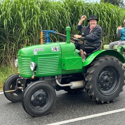 Ein Mann fährt einen grünen Traktor mit 'KLJD' vorne und lächelt, während eine andere Person hinter ihm sitzt. Sie sind von hohem Gras umgeben.