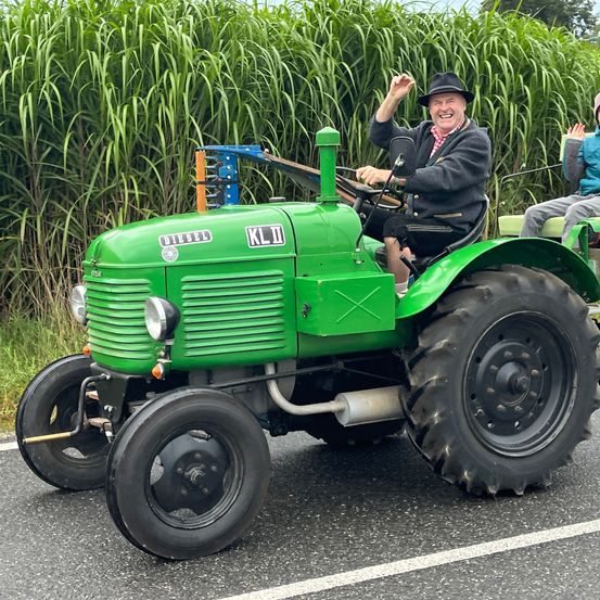 Ein Mann fährt einen grünen Traktor mit 'KLJD' vorne und lächelt, während eine andere Person hinter ihm sitzt. Sie sind von hohem Gras umgeben.