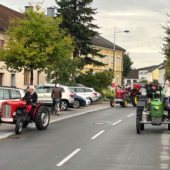 Menschen fahren mit Traktoren die Straße entlang. Eine Frau auf einem roten Traktor führt. Eine Gruppe von Menschen auf grünen und roten Traktoren folgt.