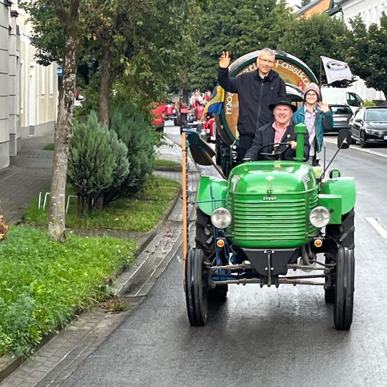 Ein grüner Traktor fährt auf einer Straße, mit zwei Personen darin. Einer winkt und der andere lächelt. Der Traktor hat einen Fass am Heck. Es gibt Büsche und einen Baum an der Seite.