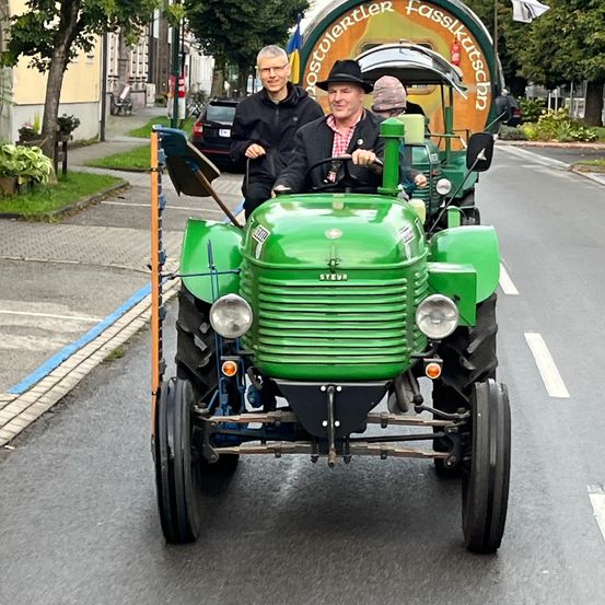 Zwei Männer in einem grünen Traktor fahren die Straße hinunter, mit einem Fass an der Rückseite. Bäume und geparkte Autos sind im Hintergrund.