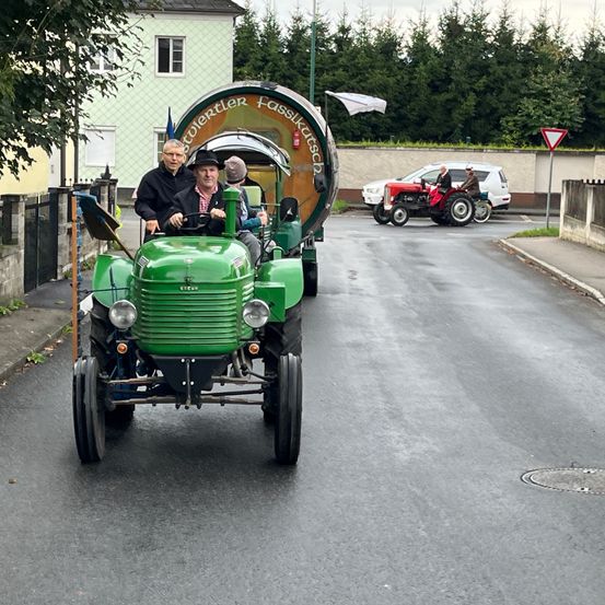 Ein grüner Traktor mit einem Fass fährt auf einer Straße. Dahinter fährt ein weiterer Traktor mit zwei darauf sitzenden Personen. Bäume und ein Gebäude sind im Hintergrund.