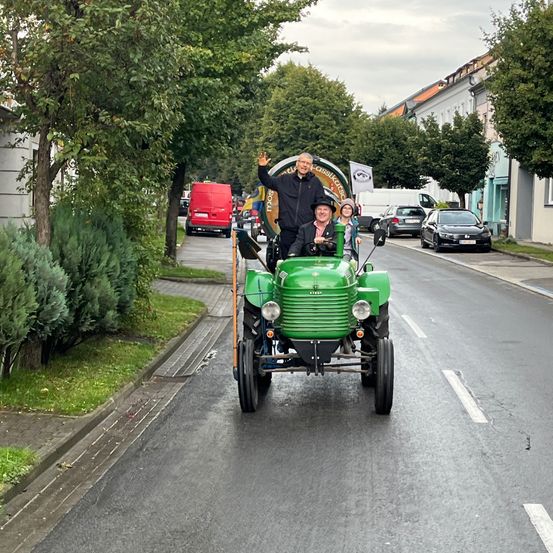 Zwei Männer fahren mit einem grünen Traktor die Straße entlang und winken zur Kamera. Bäume und geparkte Autos säumen die Straße.