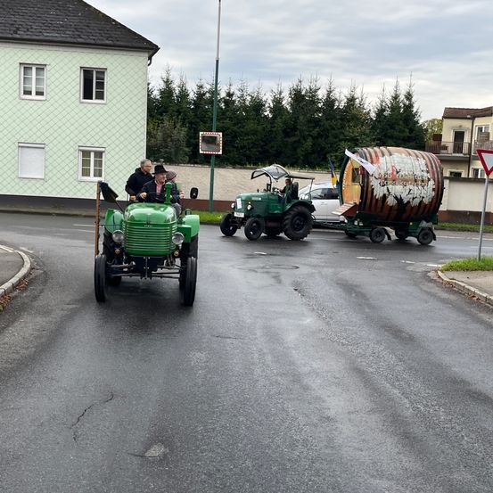 Ein grüner Traktor mit einem Fass-Anhänger fährt die Straße entlang, gefolgt von einem weiteren Traktor und einem Auto. Im Hintergrund befindet sich ein Gebäude mit Balkon.