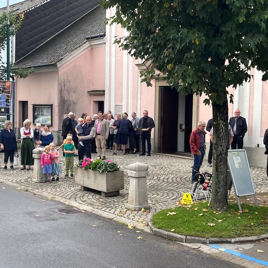 Menschen versammeln sich vor einem rosa Gebäude, wahrscheinlich einer Kirche, mit einem Baum und einem Blumenbeet im Vordergrund. Ein Mann schiebt ein Fahrrad unter den Baum.