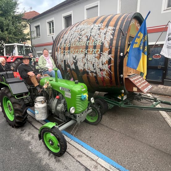 Zwei Personen sitzen auf einem Traktor, der einen großen Fass mit der Aufschrift 'Postler' zieht. An dem Fass ist eine Fahne befestigt. Der Traktor steht auf der Straße mit einem Gebäude und geparkten Fahrzeugen im Hintergrund.