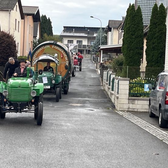 Eine Gruppe von Menschen in alten grünen Traktoren fährt eine Straße hinunter, gesäumt von Häusern, Bäumen und einem geparkten Auto.