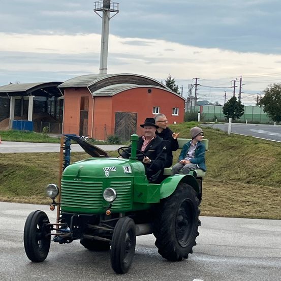 Drei Personen fahren mit einem grünen Traktor die Straße entlang. Dahinter sind ein rotes Scheunengebäude und ein Turm zu sehen.