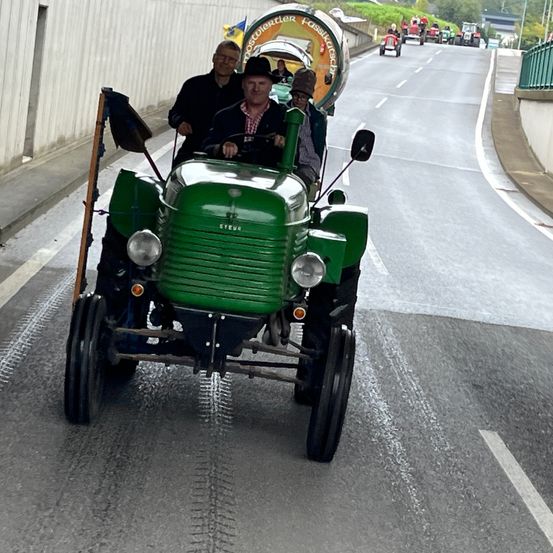 Drei Personen fahren in einem grünen Traktor auf einer Straße, gefolgt von einem weiteren Traktor. In der Ferne sind weitere Traktoren zu sehen, und die Straße wird von einer grünen Umzäunung gesäumt.