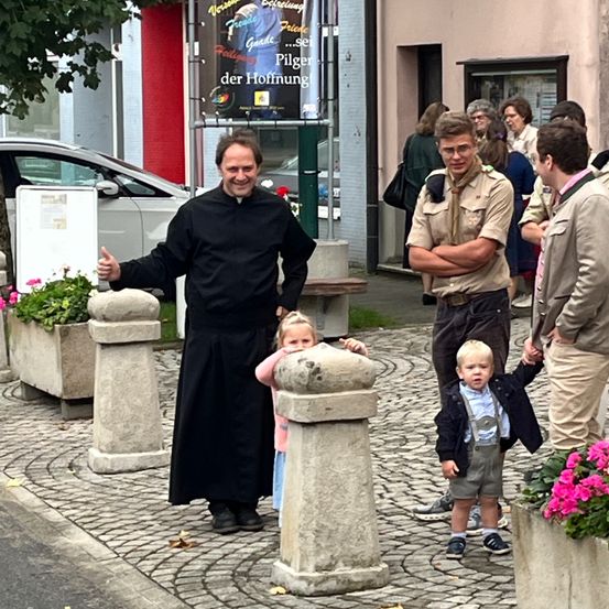 Ein Priester mit Daumen hoch steht neben einem jungen Mädchen mit einem Betonpfosten. In der Nähe werden ein Junge in Hosenträger und ein Junge in einem Pfadfinderuniform von einem Erwachsenen gehalten. Dahinter unterhält sich eine Frau mit einer anderen Person.