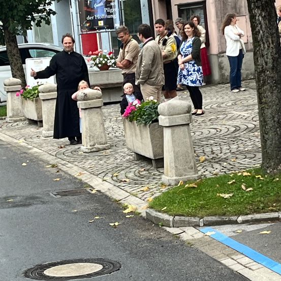 Ein Priester winkt mit mehreren Kindern und Erwachsenen auf einem Bürgersteig mit Pflanzen und einem Baum. Ein Gebäude mit einem Schild befindet sich hinter ihnen.