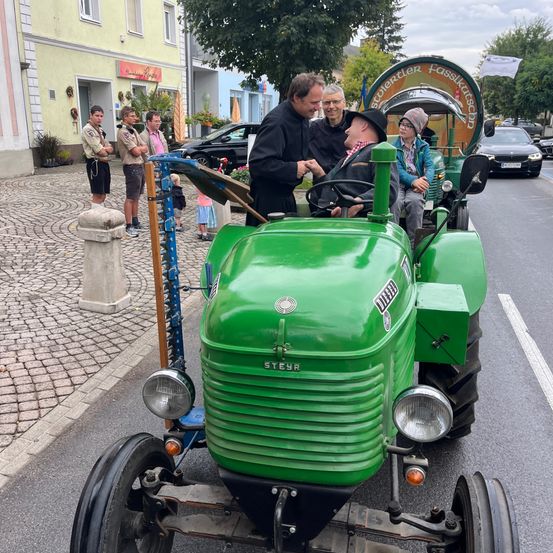 Eine Gruppe von Menschen steht auf einer Kopfsteinpflasterstraße. Ein Mann in einem schwarzen Mantel spricht mit einem Mann in einem Hut und einer Frau mit Brille. Sie sind in einem grünen Traktor mit dem Wort STEYR auf der Vorderseite.