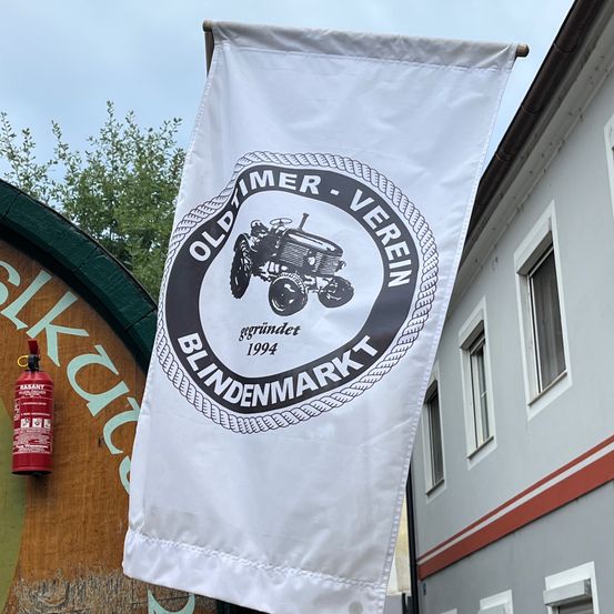 Eine weiße Flagge mit einem Traktorlogo, dem Text 'OLDTIMER VEREIN' und dem Jahr '1994'. Die Fahne hängt neben einem Gebäude mit einem roten Feuerlöscher auf der linken Seite.