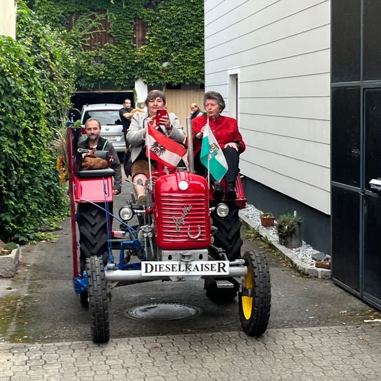Drei Personen fahren auf einem roten Traktor, zwei vorne und eine hinten. Sie halten Flaggen, und ein Mann geht hinter ihnen. In der Nähe steht ein Gebäude mit weißer Verkleidung und einem geparkten Auto.