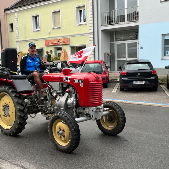 Ein Mann fährt einen roten Traktor auf der Straße, hinter ihm weht eine Flagge. Dahinter sind mehrere Autos geparkt.