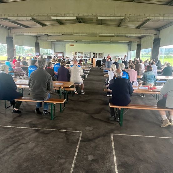 Innenbereich mit Esstischen, an denen Menschen sitzen, einer Bühne mit einer sprechenden Person und großen Fenstern mit Blick nach draußen.