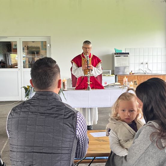 Ein Priester in roter und weißer Kleidung führt eine Zeremonie mit einem Kreuz durch, während eine Familie, einschließlich eines kleinen Mädchens, von vorne zuschaut.