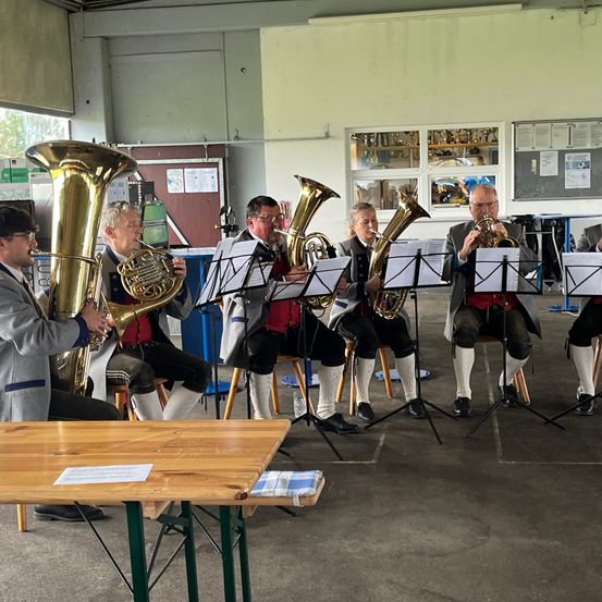 Eine Gruppe von Musikern spielt Blechblasinstrumente in einem Raum mit einem Holztisch. Sie sitzen auf Hockern und sind in traditioneller Kleidung gekleidet.