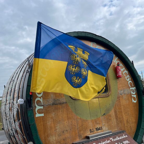 Ein großer Fass zeigt eine blau-gelbe Flagge mit einem Schlosswappen. Ein Feuerlöscher ist am Fass montiert. Das Fass trägt die Aufschrift 'BJ. 1945'.