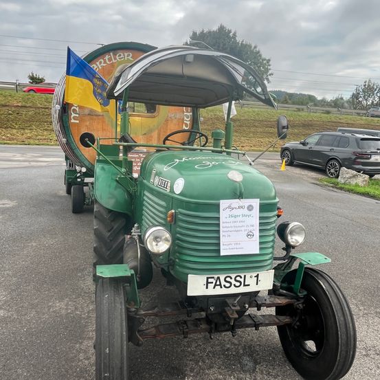 Ein grüner Oldtimer-Traktor mit einem Fass auf dem Anhänger und einer blau-gelben Flagge steht auf der Straße. Ein Auto ist am Straßenrand geparkt.