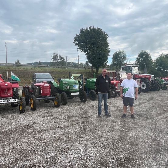 Zwei Männer stehen vor einer Reihe von Oldtimer-Traktoren auf einem Kiesfeld, mit einem bewölkten Himmel im Hintergrund.